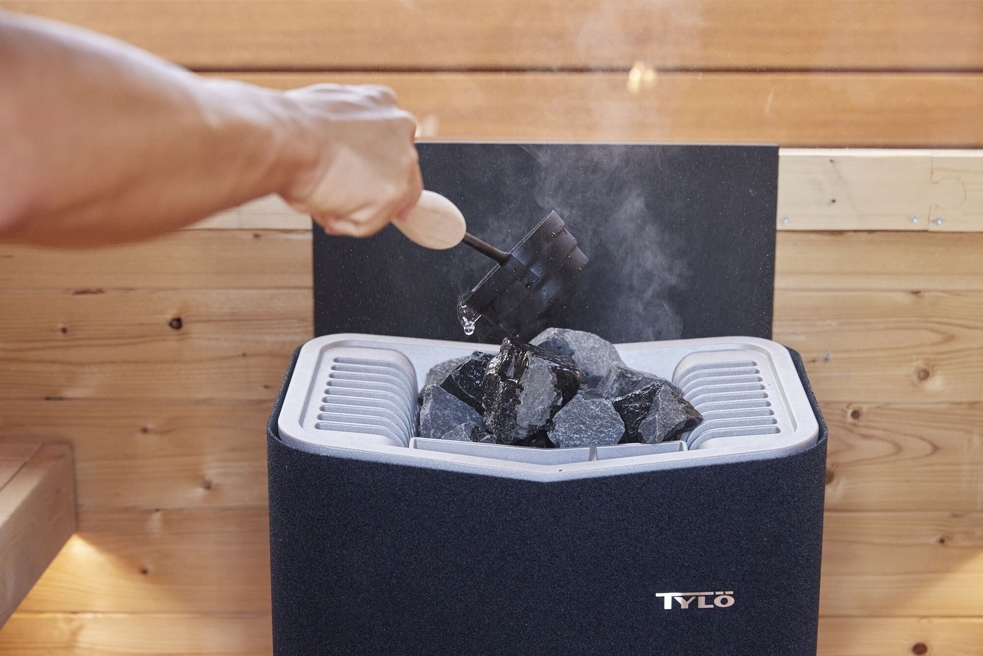 Een hand houdt een hamer boven een kolengrill, met de merknaam "TYLO" zichtbaar op de grill.