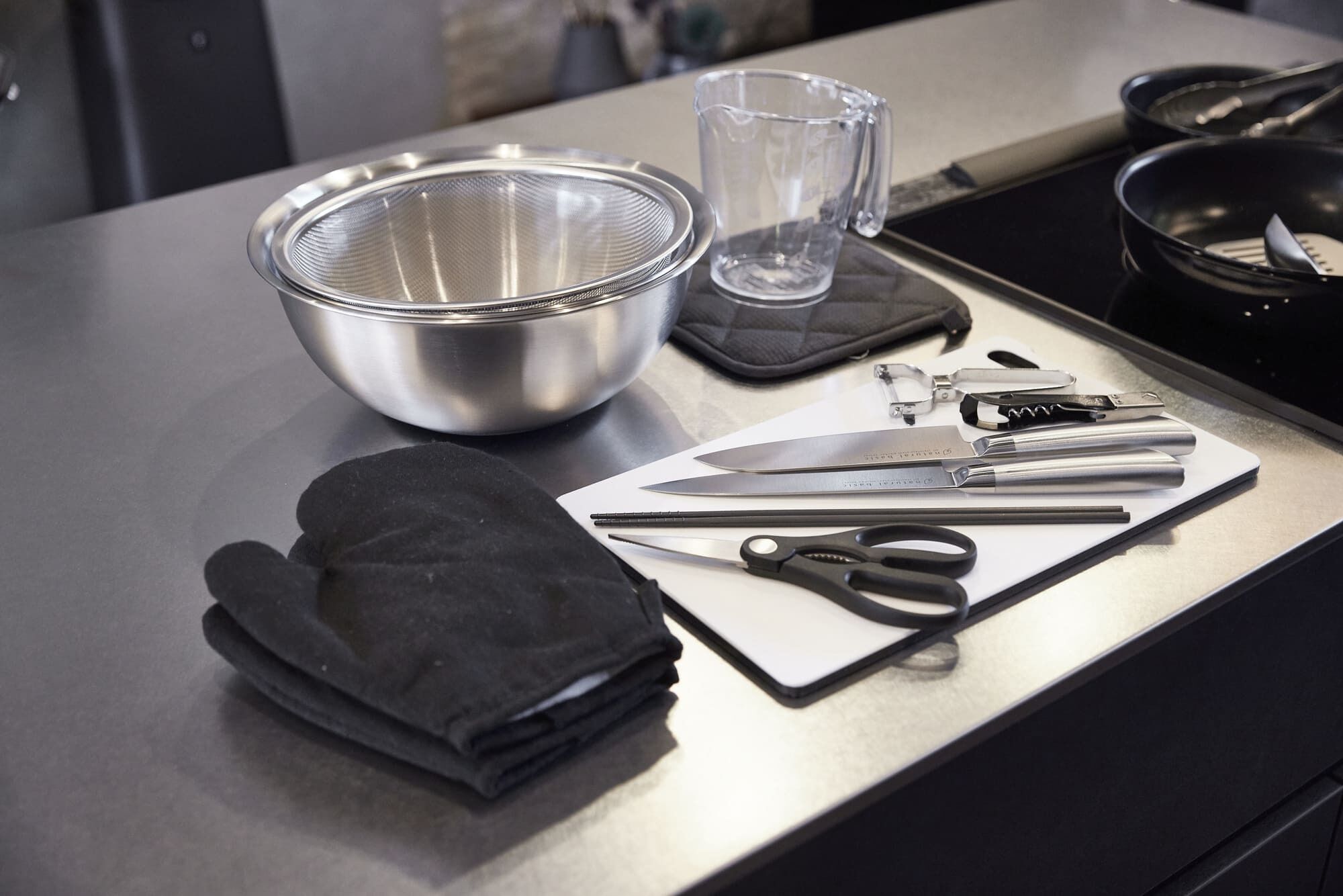Una encimera de cocina con un bol de acero inoxidable, una jarra de vidrio, un cuchillo, tijeras y guantes de horno negros.