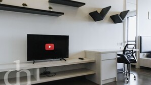 Έν α σύγχρονο σαλόνι με τηλεόραση επίπεδης οθόνης, ένα λευκό γραφείο, μια μαύρη καρέκλα γραφείου και εντοιχισμένα ράφια.
