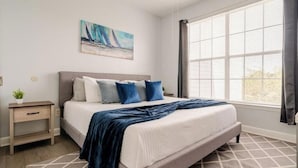 Un dormitorio con cama, mesita de noche, ventana con persianas y un cuadro en la pared.
