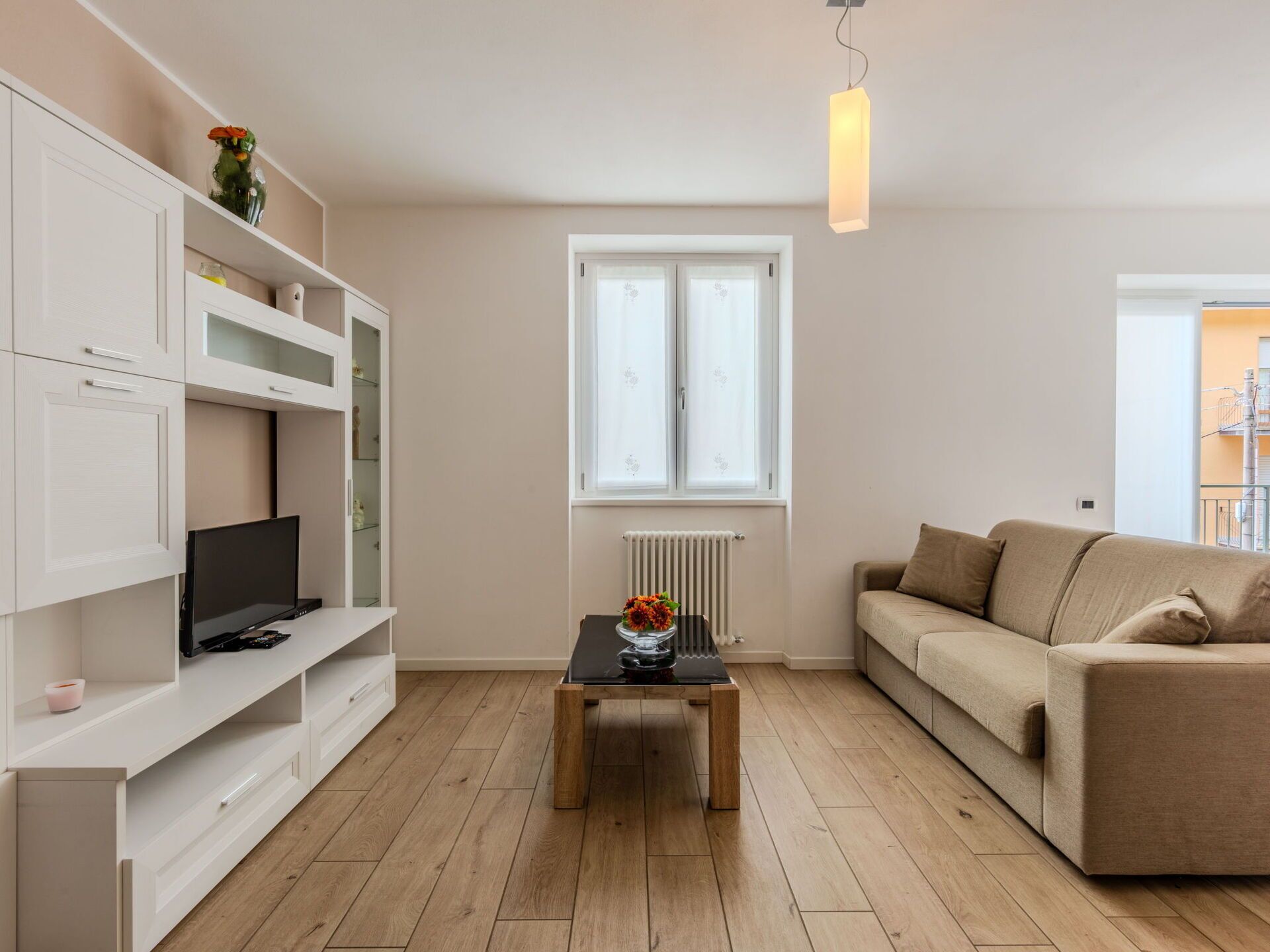 A modern living room with a beige sofa, a wooden coffee table, and a TV on a white cabinet. A window with a radiator is visible, and a hanging light fixture is above the table.