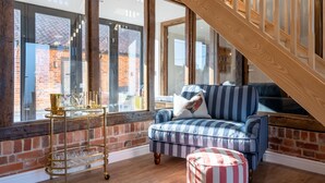 Una sala de estar acogedora con un sofá azul a rayas, una mesa de centro de vidrio y un reposapiés a rayas. Una escalera de madera conduce a un nivel superior.