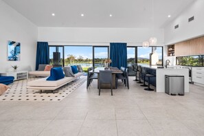 Una sala de estar moderna con sofá, mesa de comedor y sillas, una cocina con pileta y alacenas, y grandes ventanas con vista al exterior.