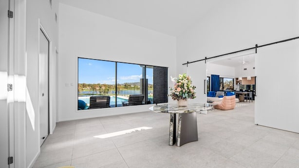 A spacious modern living area with a large window overlooking a pool and landscape, a round glass table with a floral centerpiece, and a minimalist design. - Luxury Lakefront Getaway | 6BR Home w/ Pool & Cinema (Tamborine)