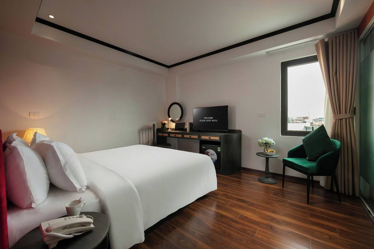 A hotel room with a bed, a TV, a small table with a flower vase, a green armchair, and a window with curtains.