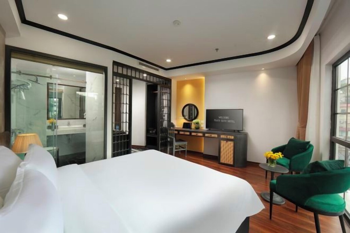 Une chambre d’hôtel moderne avec un grand lit, un bureau avec une télévision et un coin salon avec des fauteuils verts.