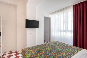 Un dormitorio con piso a cuadros, una cama con colcha floral colorida, un televisor montado y una ventana con cortinas traslúcidas y un drape rojo.