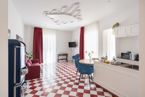 A room with a checkered floor, a dining area with blue chairs, a red sofa, a kitchen with white cabinets, and a vintage blue refrigerator.