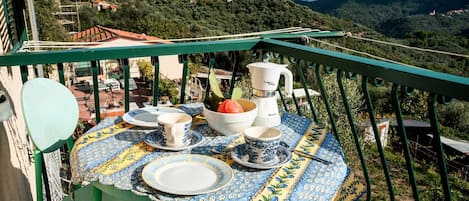 A table set with blue and yellow patterned tablecloth, white plates, blue and white cups, a bowl, and a white coffee maker on a balcony overlooking a mountainous landscape.