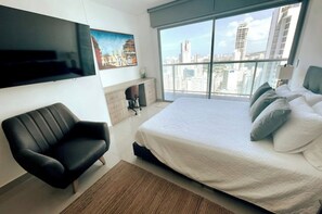 A modern hotel room with a large bed, a black leather chair, a flat-screen TV mounted on the wall, and a city view through large windows. - Panoramic High-Rise Oceanview Condo in Bocagrande (Bocagrande)
