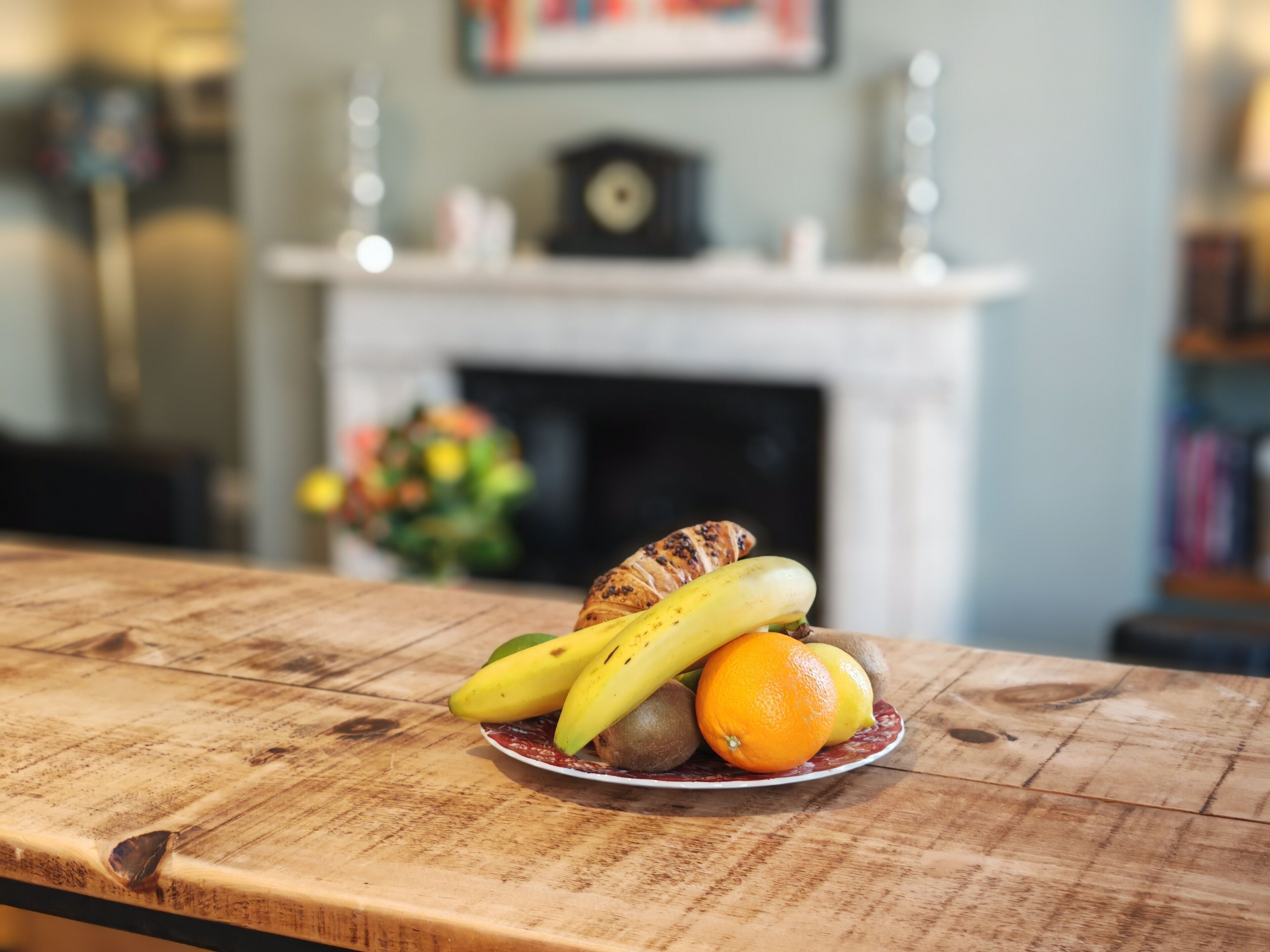 Una mesa de madera con un plato de fruta y un reloj sobre una repisa al fondo.