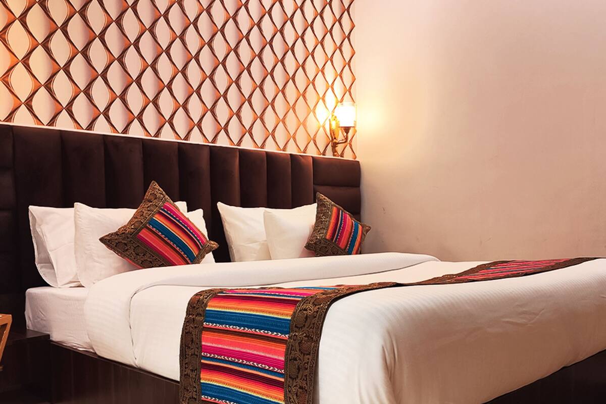 Una cama bien hecha con sábanas blancas y almohadas de colores, un cabecero oscuro y un papel tapiz estampado al fondo.