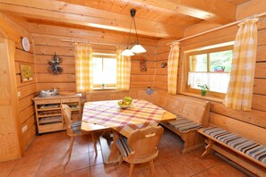 A wooden cabin interior with a dining table, benches, a clock, and a window with curtains.