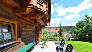 Une cabane en bois avec une table et des chaises à l’extérieur, un banc, et une vue sur un paysage verdoyant avec des maisons en arrière-plan.