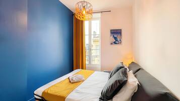 A hotel room with a bed, a hanging light fixture, a window with curtains, and a poster on the wall.