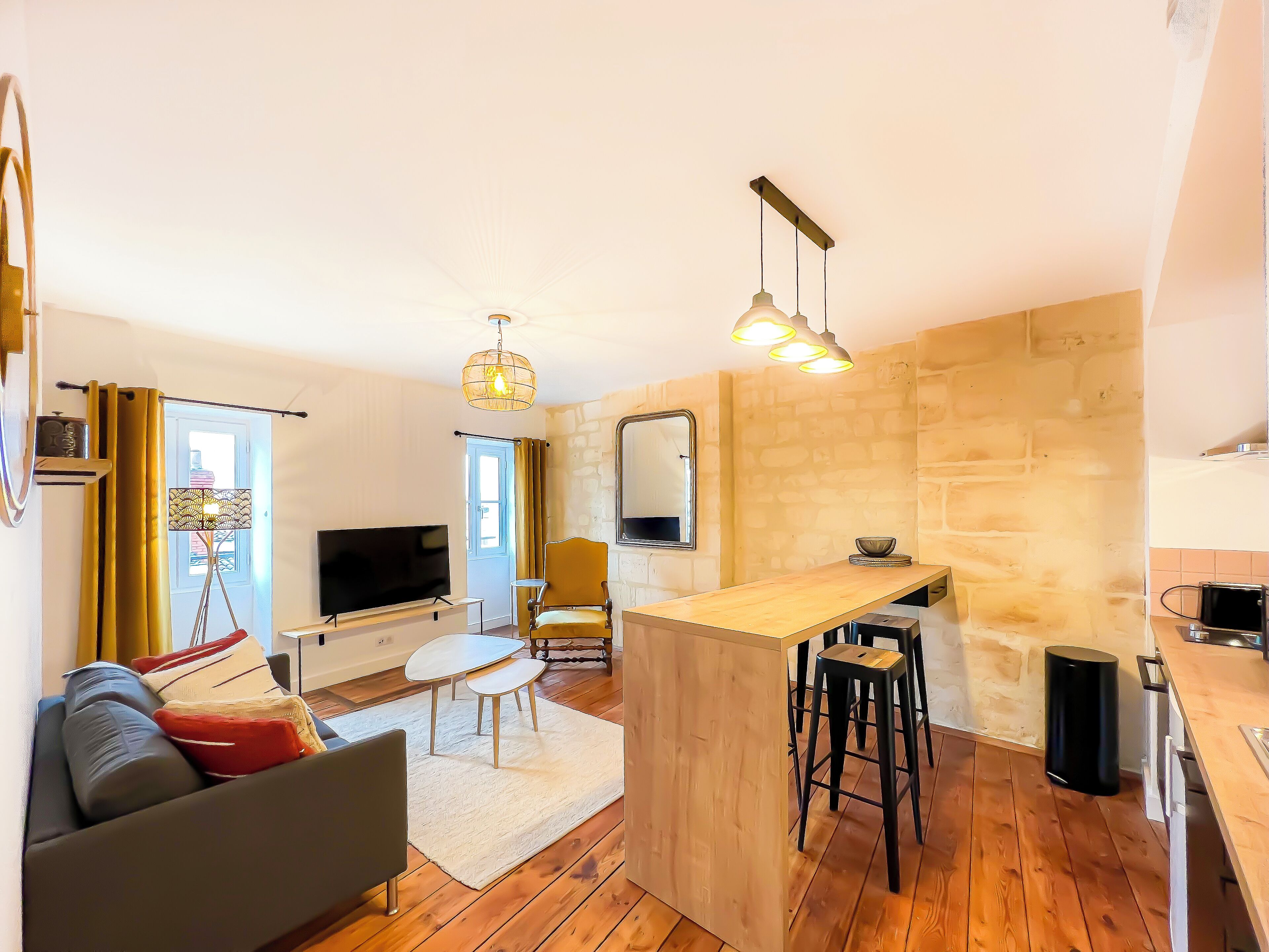A modern living room with a flat-screen TV, a sofa, a dining area with stools, and a kitchen area with a wooden counter and bar stools.