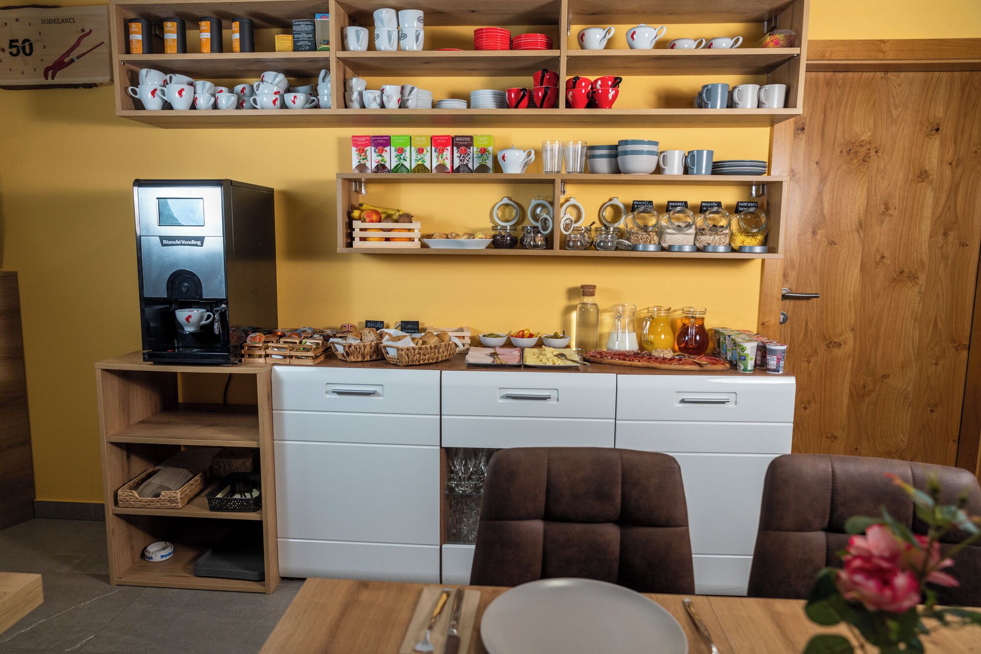 A kitchenette with a coffee machine, shelves with kitchenware, and a variety of food items.