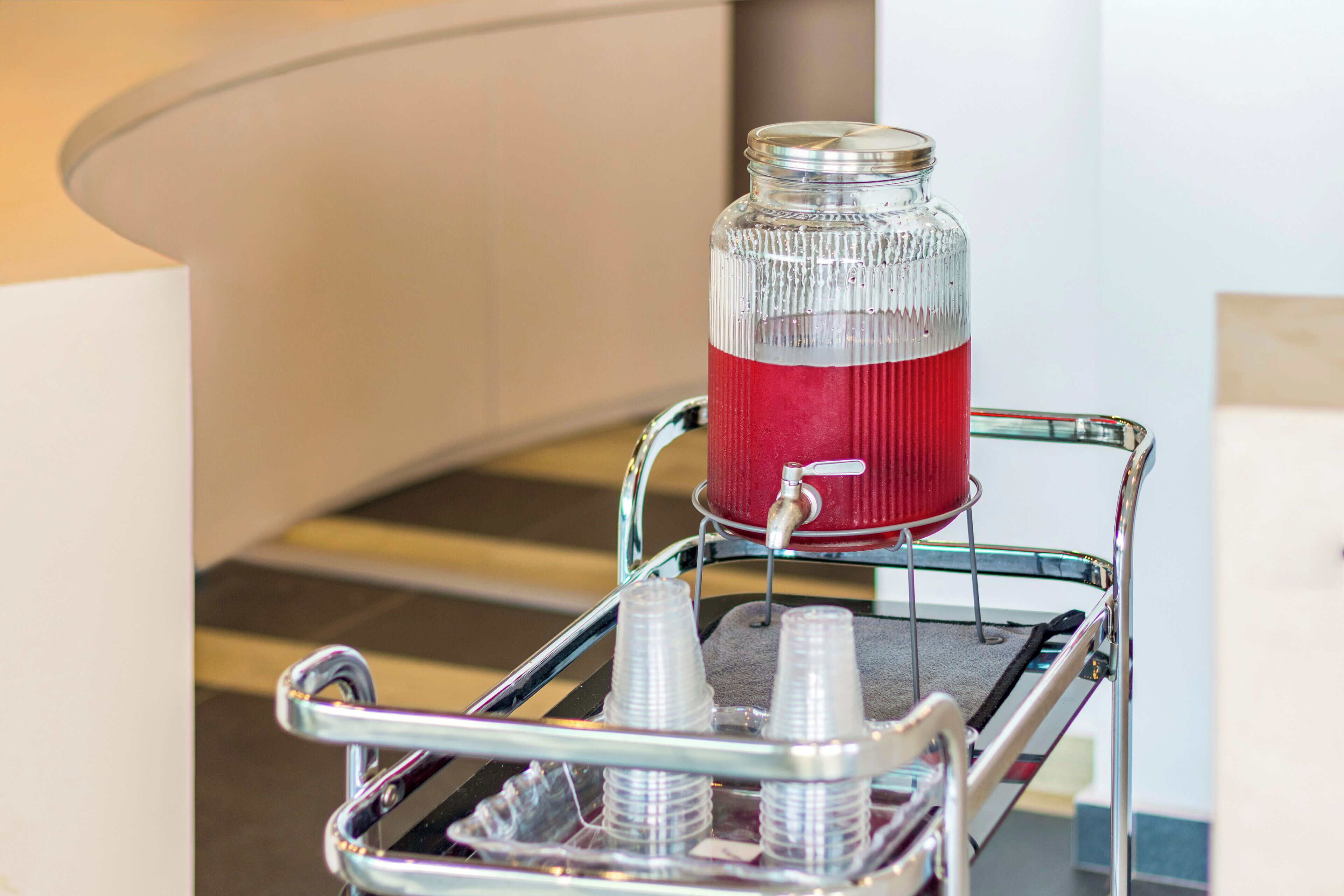 A beverage cart with a glass jar filled with a red drink, a water dispenser, and several plastic cups.