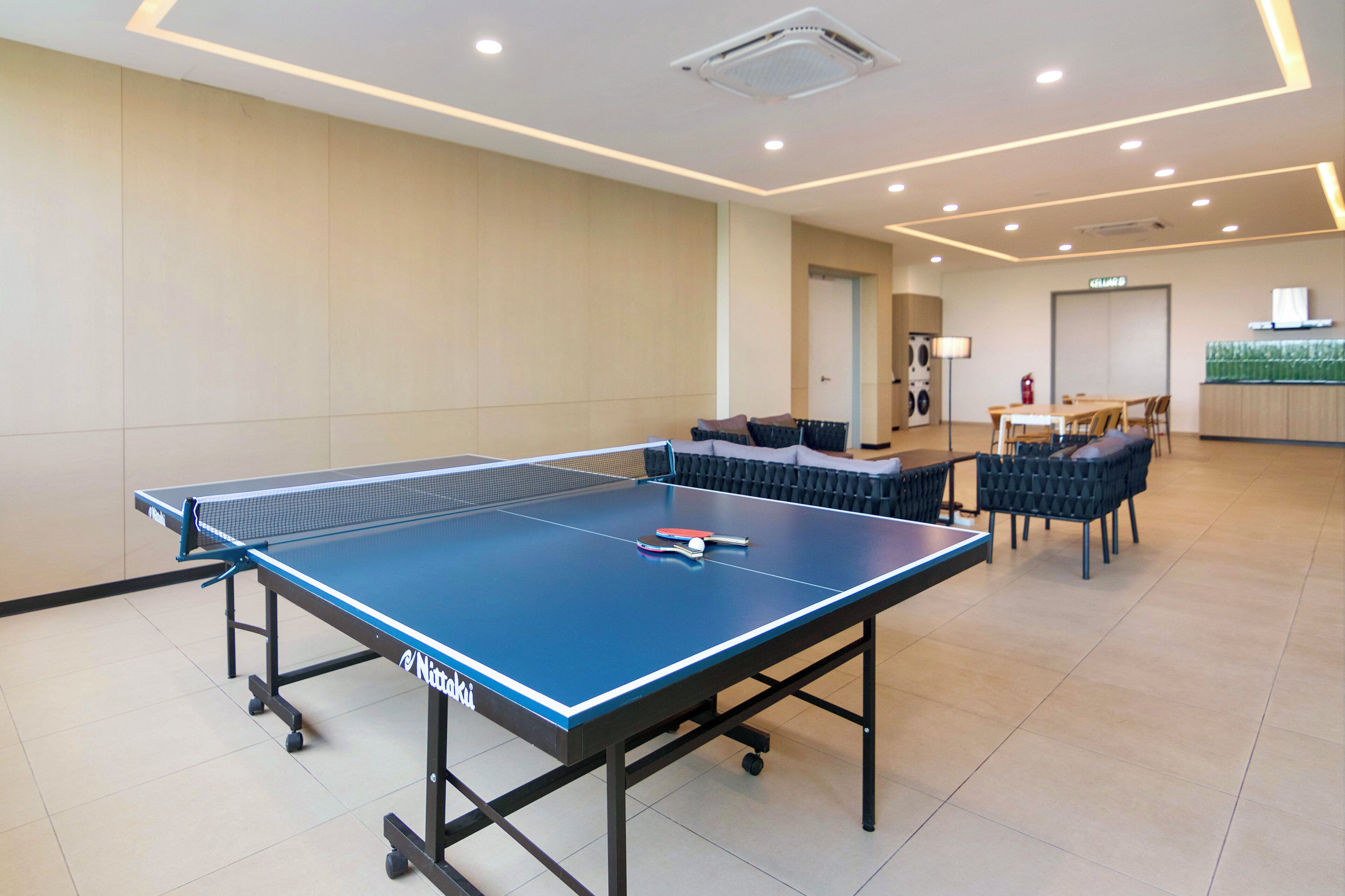 Une table de ping-pong avec une raquette et une balle de tennis de table, entourée de chaises et d’un canapé, dans une pièce bien éclairée avec un sol carrelé et une porte visible au fond.