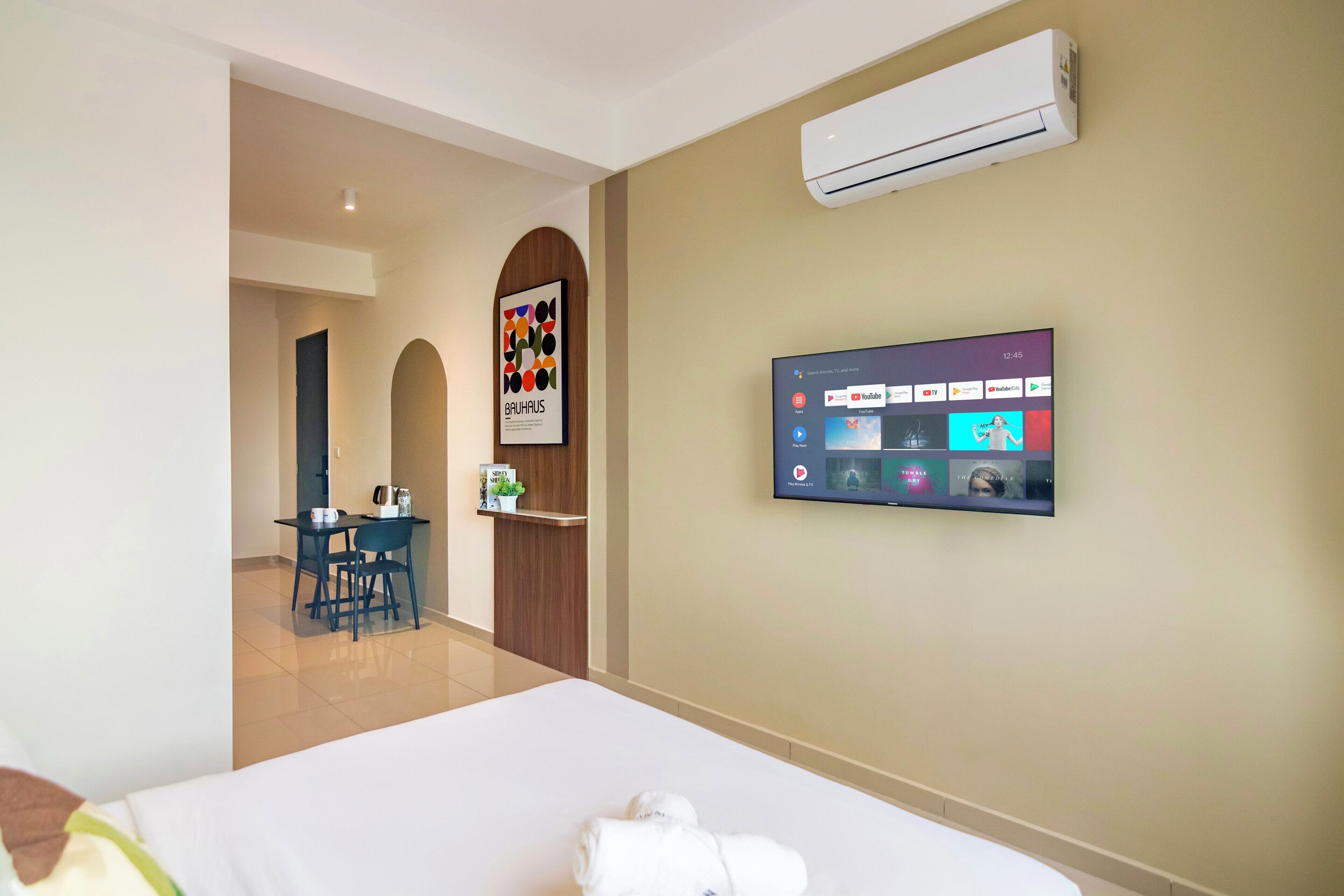 Habitación de hotel con cama, televisor montado en la pared que muestra varios canales, una pequeña mesa con sillas y una zona de kitchenette.