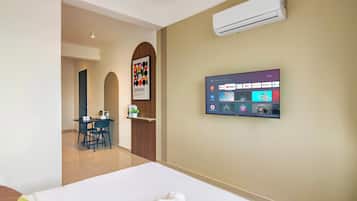 Habitación de hotel con cama, televisor montado en la pared que muestra varios canales, una pequeña mesa con sillas y una zona de kitchenette.