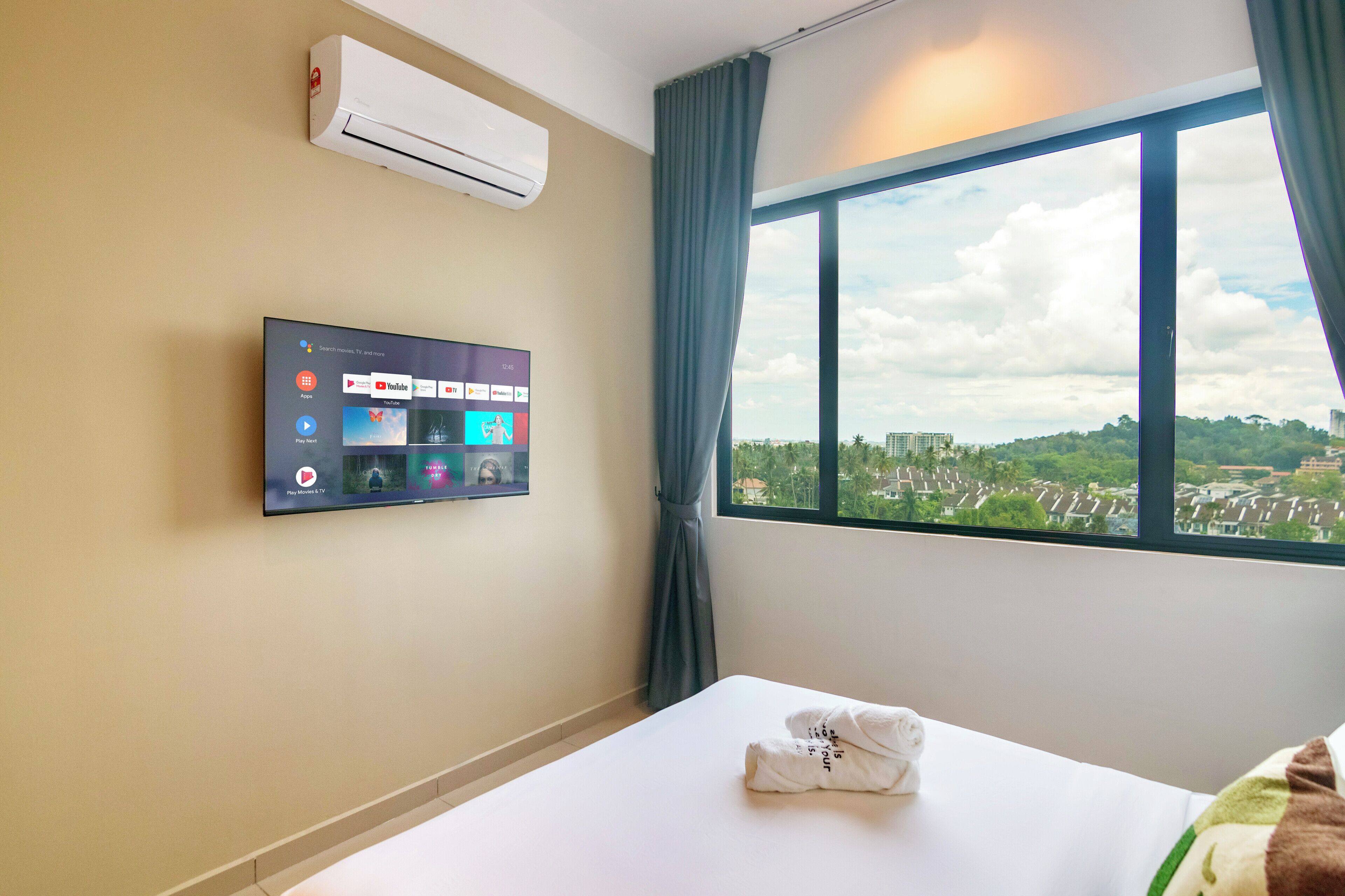 A hotel room with a bed, a flat-screen TV mounted on the wall displaying various streaming services, an air conditioning unit, and a large window offering a city view.