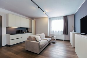 A modern living space with a sofa, a small side table, a flat-screen TV, and a kitchen area. - Ah Virgilio Apartments Resort (Mantua)