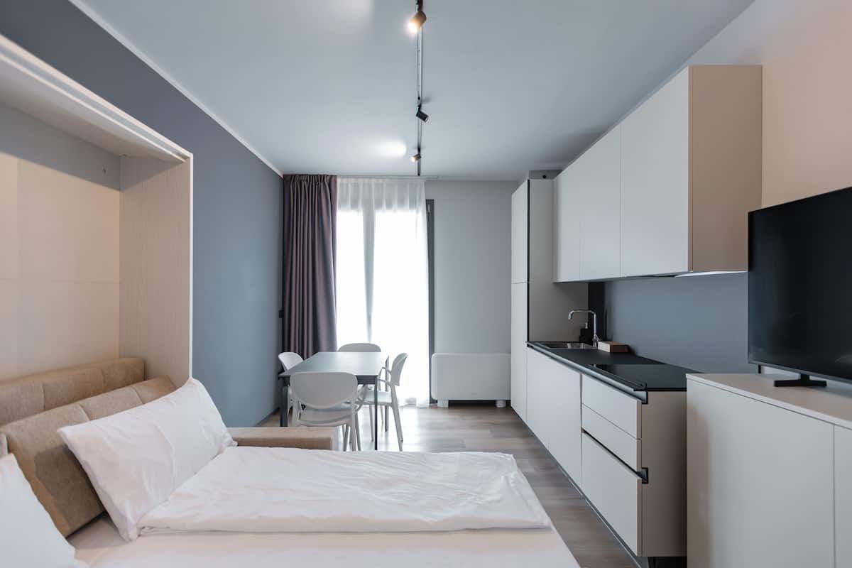 A modern hotel room with a bed, a dining table, and a kitchen area.