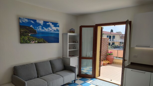 Un salon avec un canapé gris, une table à manger avec des chaises, un meuble blanc, un tableau représentant un paysage côtier et une porte-fenêtre coulissante donnant sur un balcon extérieur agrémenté de plantes en pot.
