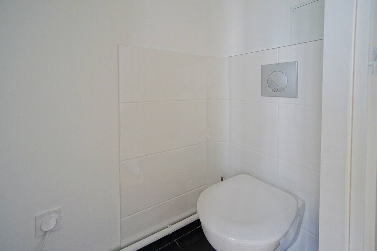 Et lille badeværelse med hvide fliser, et toilet og en vægmonteret skylleknap.