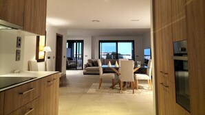 Une cuisine moderne avec des meubles en bois, un coin repas avec une table et des chaises, et un salon avec un canapé et une vue dégagée sur l’extérieur grâce à de grandes fenêtres.