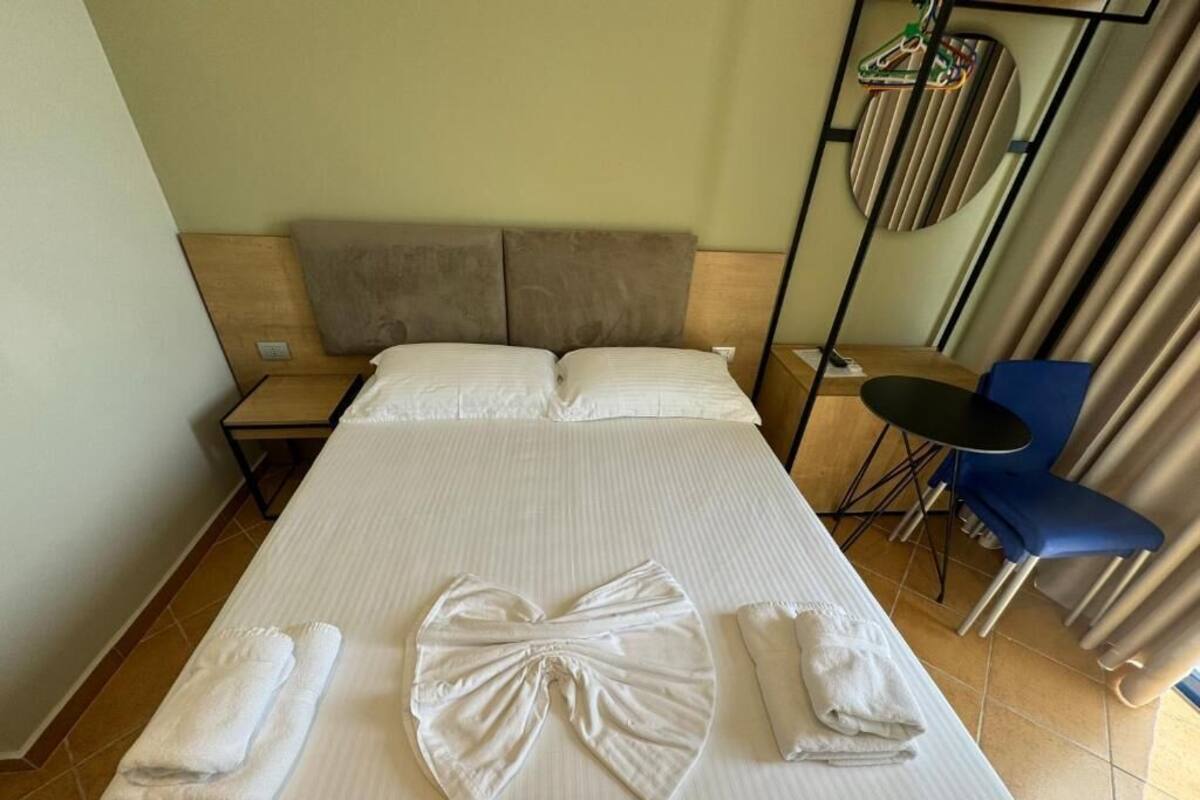 A hotel room with a large bed, two pillows, a bedside table, a chair, and a small table with a chair.