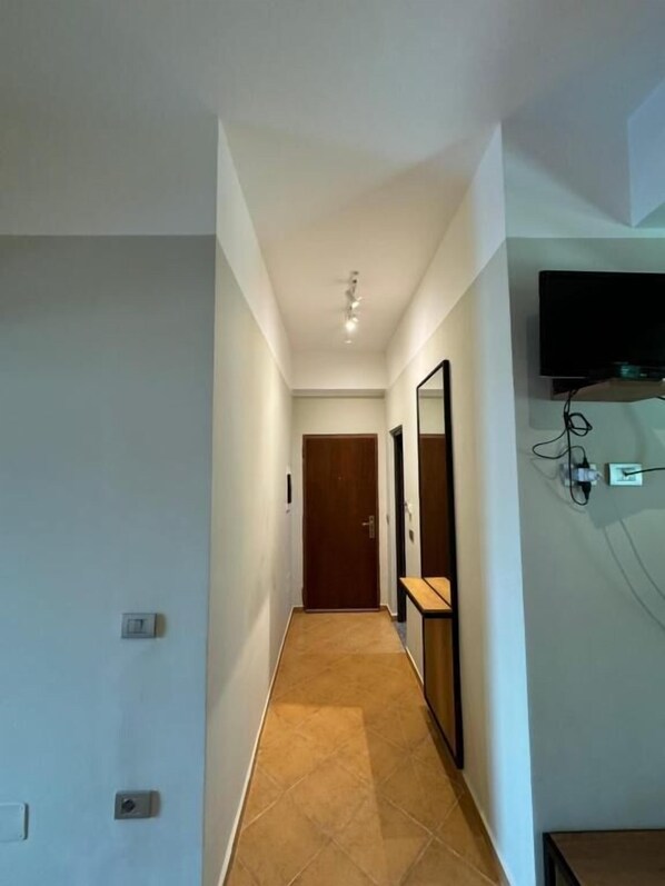 A narrow hallway with a wooden door at the end, a mirror on the right wall, and a flat-screen TV mounted above a power outlet. - Hotel Cekodhima (Orikum)