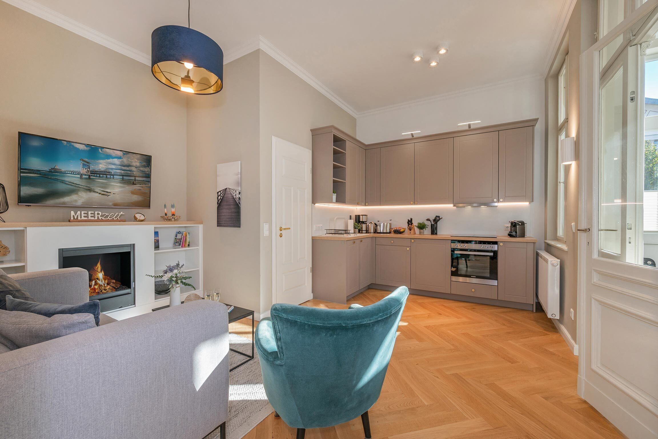 Un salon moderne avec une cheminée, un fauteuil de couleur turquoise et une cuisine équipée d’appareils électroménagers intégrés.