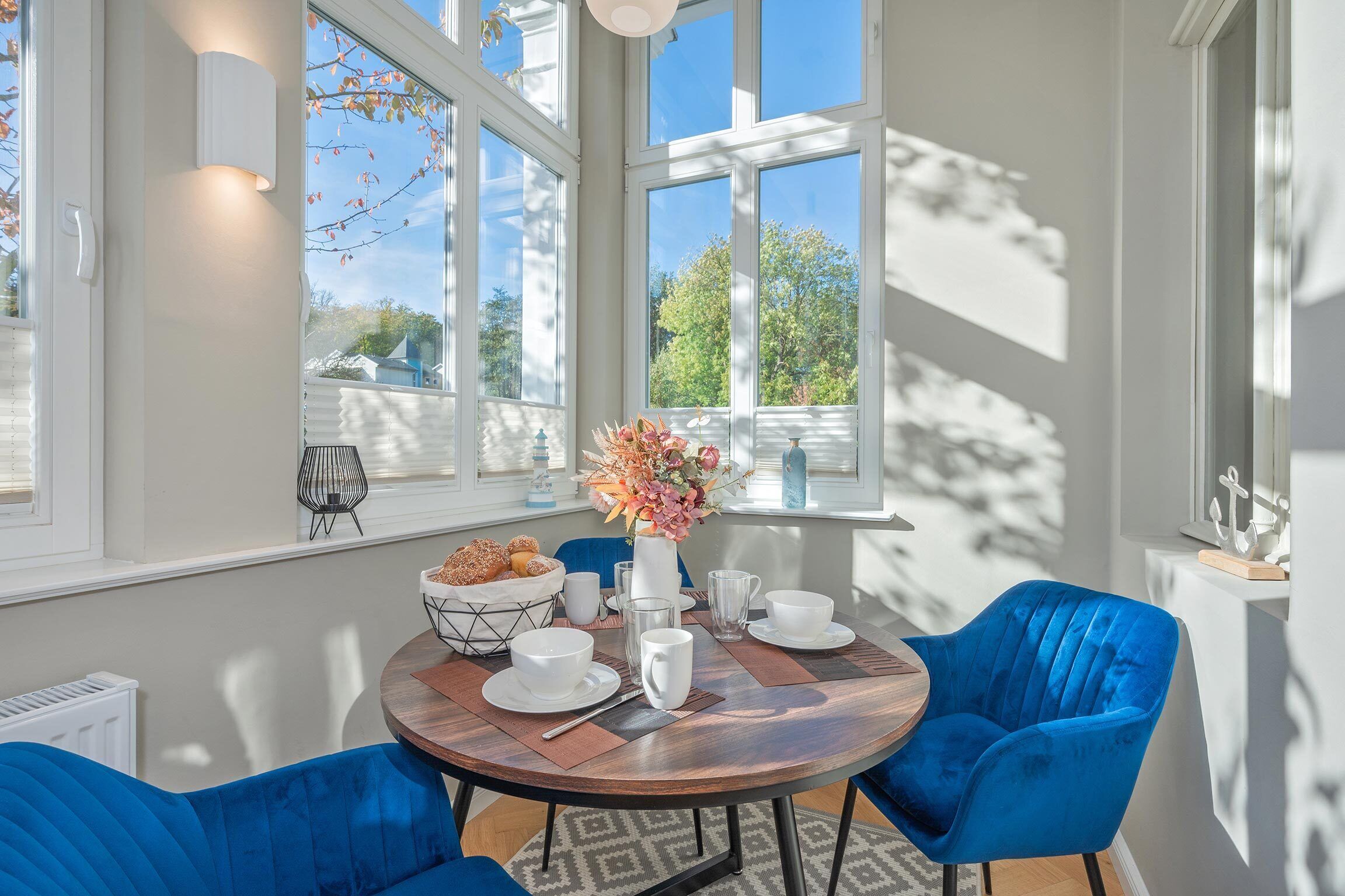 Un coin repas avec une table ronde, des chaises bleues, de la vaisselle blanche et un vase de fleurs.