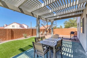 Une terrasse couverte avec une table et des chaises en bois, un barbecue, et une vue sur un mur de briques et des maisons au loin.