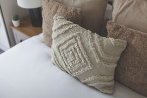 Una cama con almohadas color beige y marrón, una planta pequeña en maceta y una lámpara en una mesita de noche.