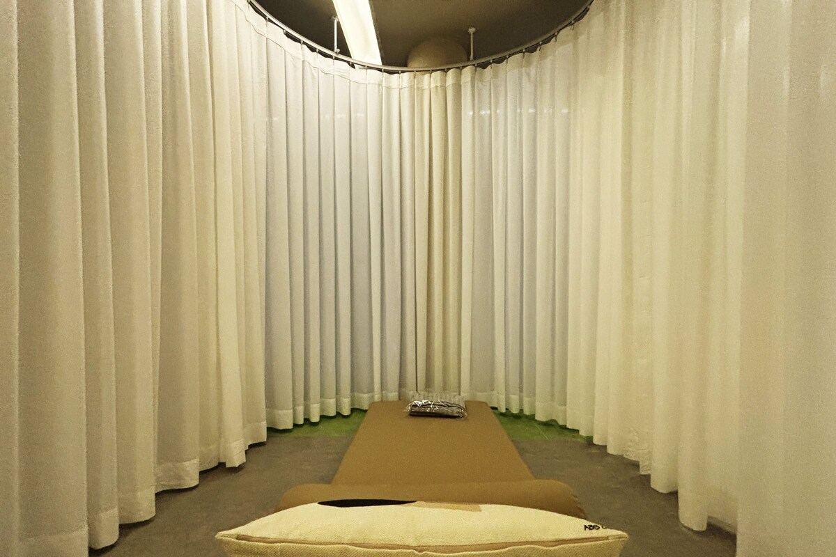 A room with a long, narrow bench and a single chair, surrounded by tall, white curtains.