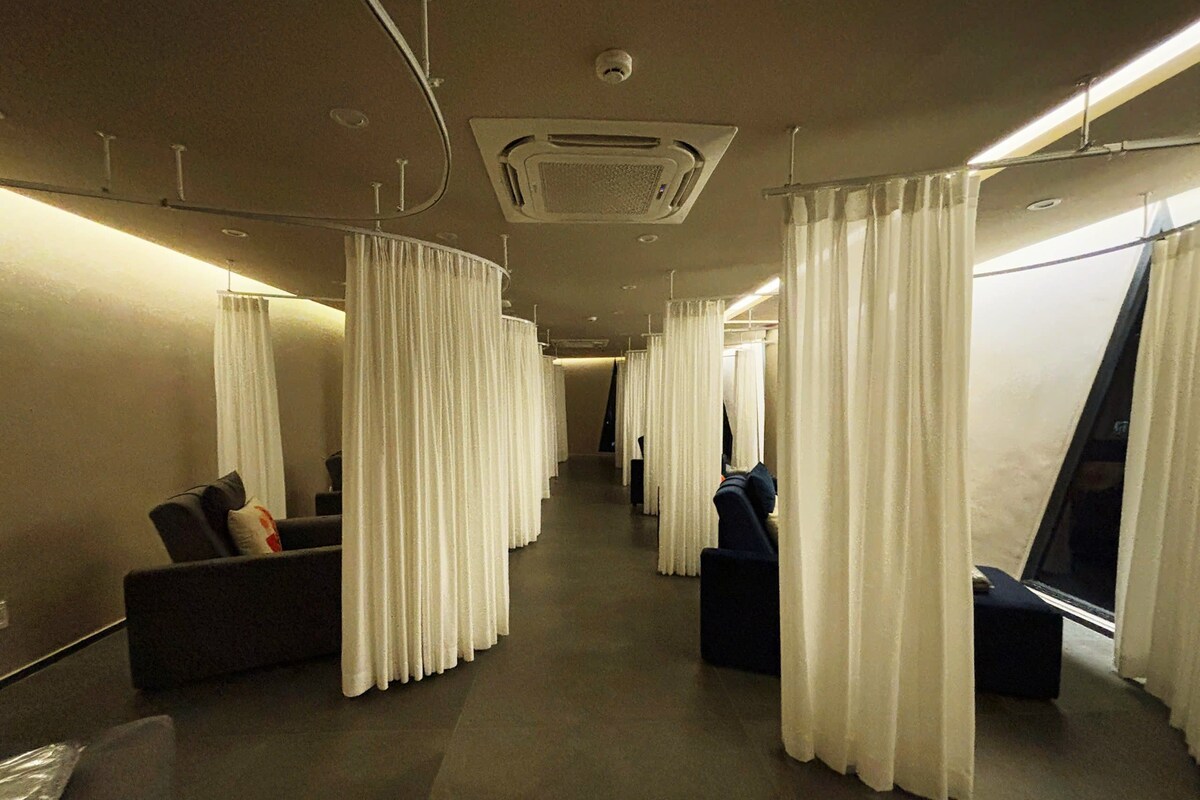 Un couloir d’hôtel bordé de rangées de coins salon séparés par des rideaux, avec un appareil de climatisation fixé au plafond et une caméra de surveillance bien visible.