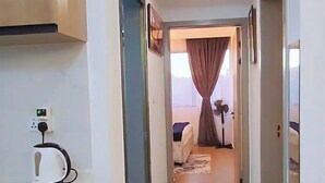 A narrow hallway with a kitchenette on the left, a bed with a nightstand and lamp in the center, and a bathroom visible through an open door. - 2 Bed Apartment Sleeps 4 - Balcony, Parking, Wifi (Nairobi)