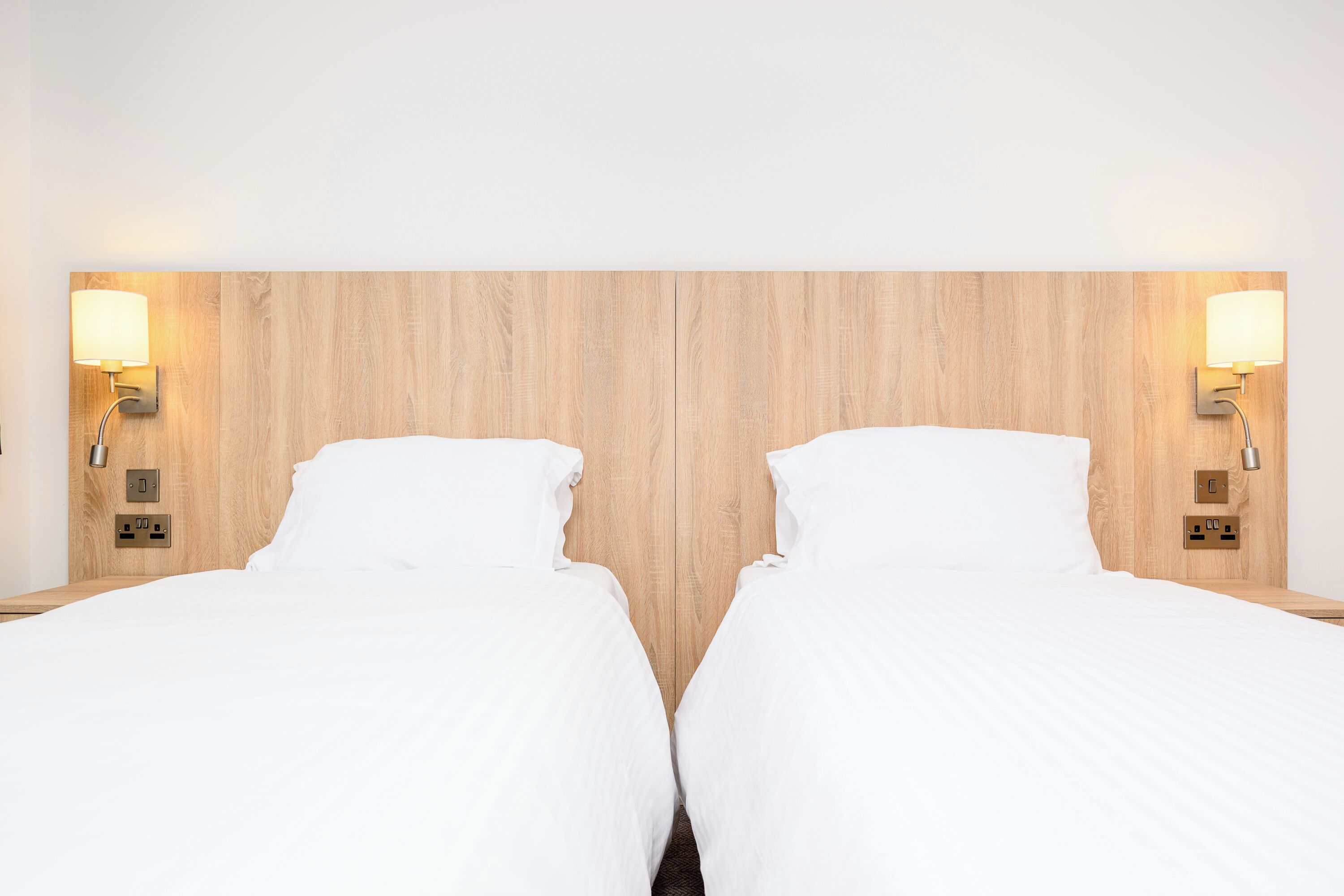 A double room with two beds, each with a white bedspread and pillows, flanked by wooden headboards and wall-mounted nightstands with lamps.