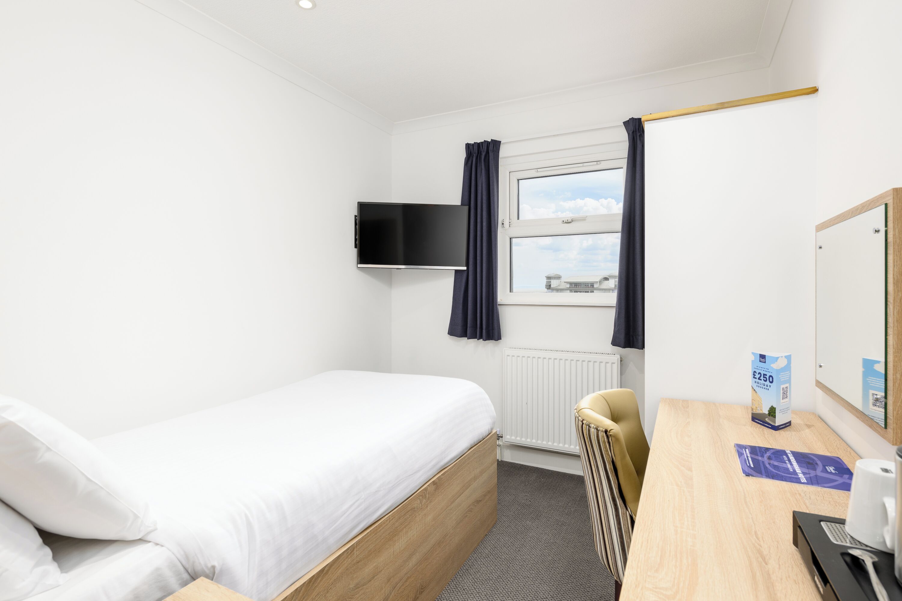 A hotel room with a single bed, a desk with a chair, a flat-screen TV mounted on the wall, a window with curtains, and a radiator.