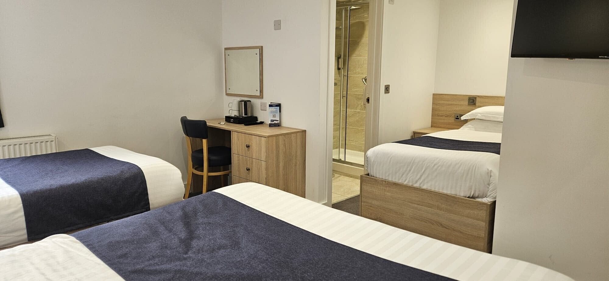 A hotel room with two beds, a desk, a chair, a mirror, and a shower area.