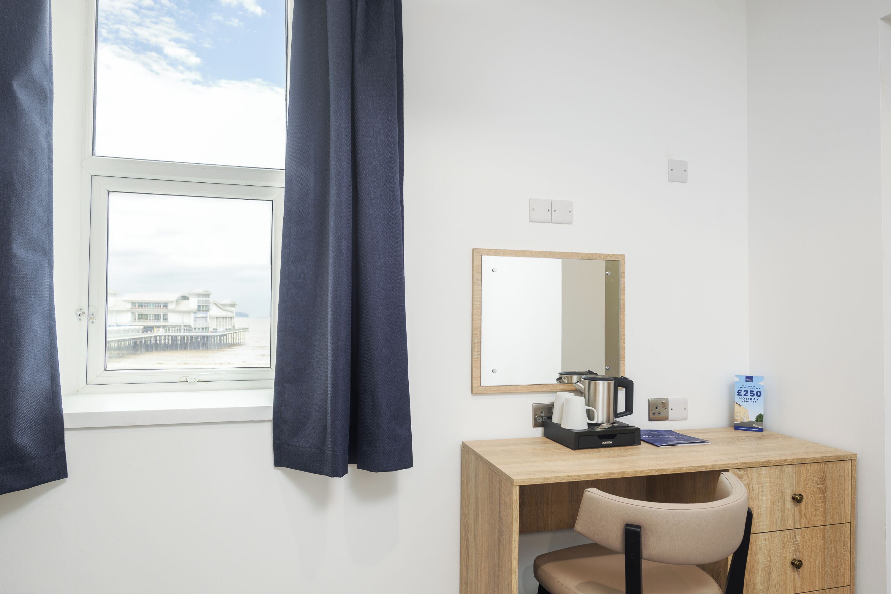 A hotel room with a wooden desk, a chair, a mirror, a window with curtains, and a view of a building outside.