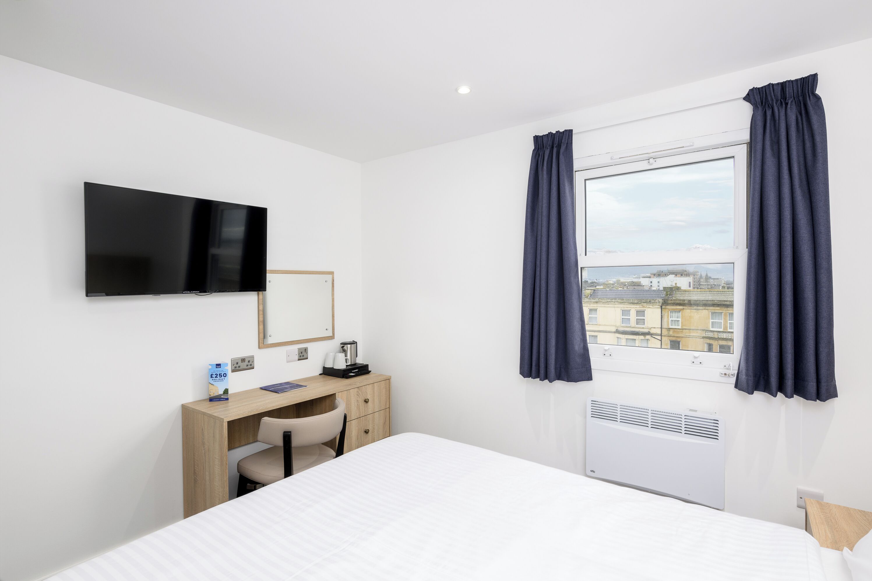 A hotel room with a bed, a desk with a chair, a flat-screen TV, a window with curtains, and a view of buildings outside.