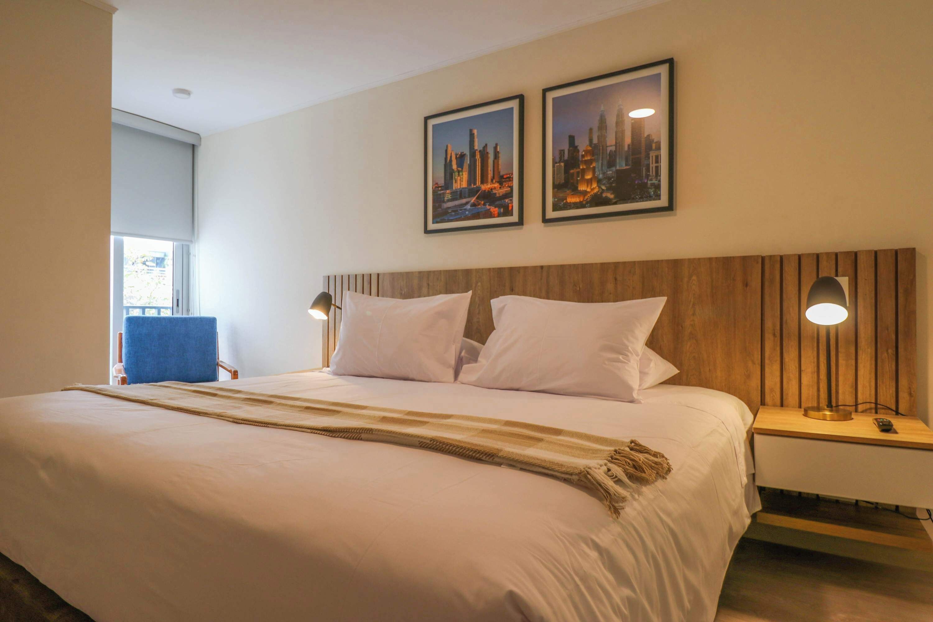 Habitación de hotel con una cama grande, dos almohadas blancas, cabecera de madera, mesita de noche con lámpara y dos cuadros en la pared.