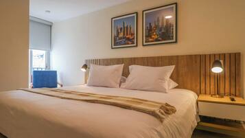 Habitación de hotel con una cama grande, dos almohadas blancas, cabecera de madera, mesita de noche con lámpara y dos cuadros en la pared.