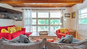 Een gezellige woonkamer met een rode bank, een grijze bank, en een rode fauteuil, een houten salontafel, en grote ramen met gordijnen.