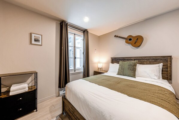 A bedroom with a bed, a guitar wall decoration, a bedside lamp, and a window with curtains. - Quebec City 2 Bedroom Treasure (Québec)