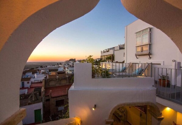Vista desde un balcón con vistas a un paisaje urbano al atardecer, mostrando un edificio blanco con un balcón y una ventana.
